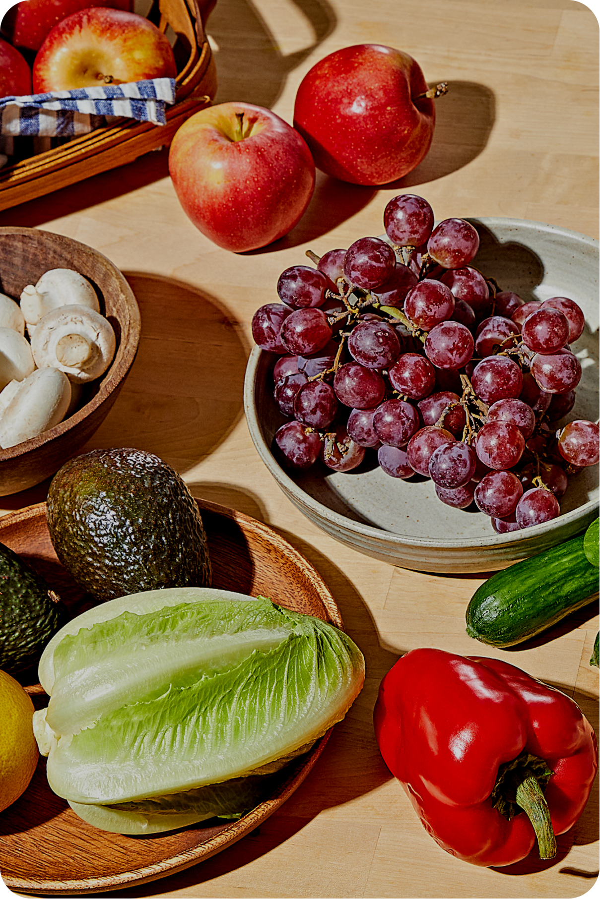 A wooden table is covered with various fresh foods. A large bunch of red grapes rests in a white bowl. Nearby are red apples, a head of romaine lettuce, an avocado, a cucumber, a red bell pepper, and a bowl of white mushrooms.