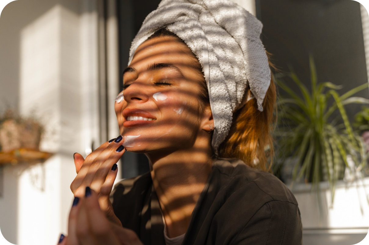 A smiling woman with a white towel wrapped around her head is applying cream to her face, illuminated by warm sunlight filtering through blinds that cast dramatic horizontal shadows across her skin.