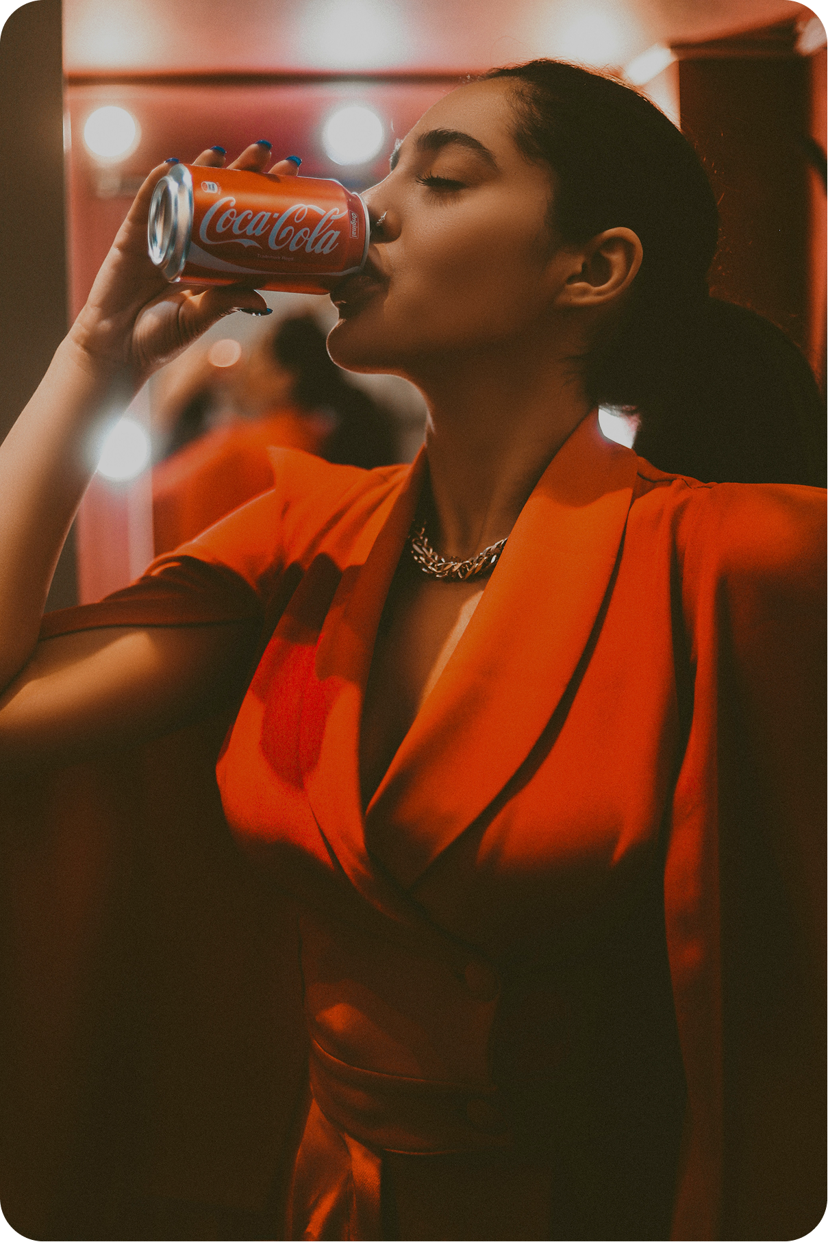 A person in a red suit jacket with a deep neckline is holding a red can of Coca-Cola to their mouth, taking a sip. Their eyes are closed, and they are wearing a silver chain necklace. The background is dimly lit with round, glowing lights behind them.