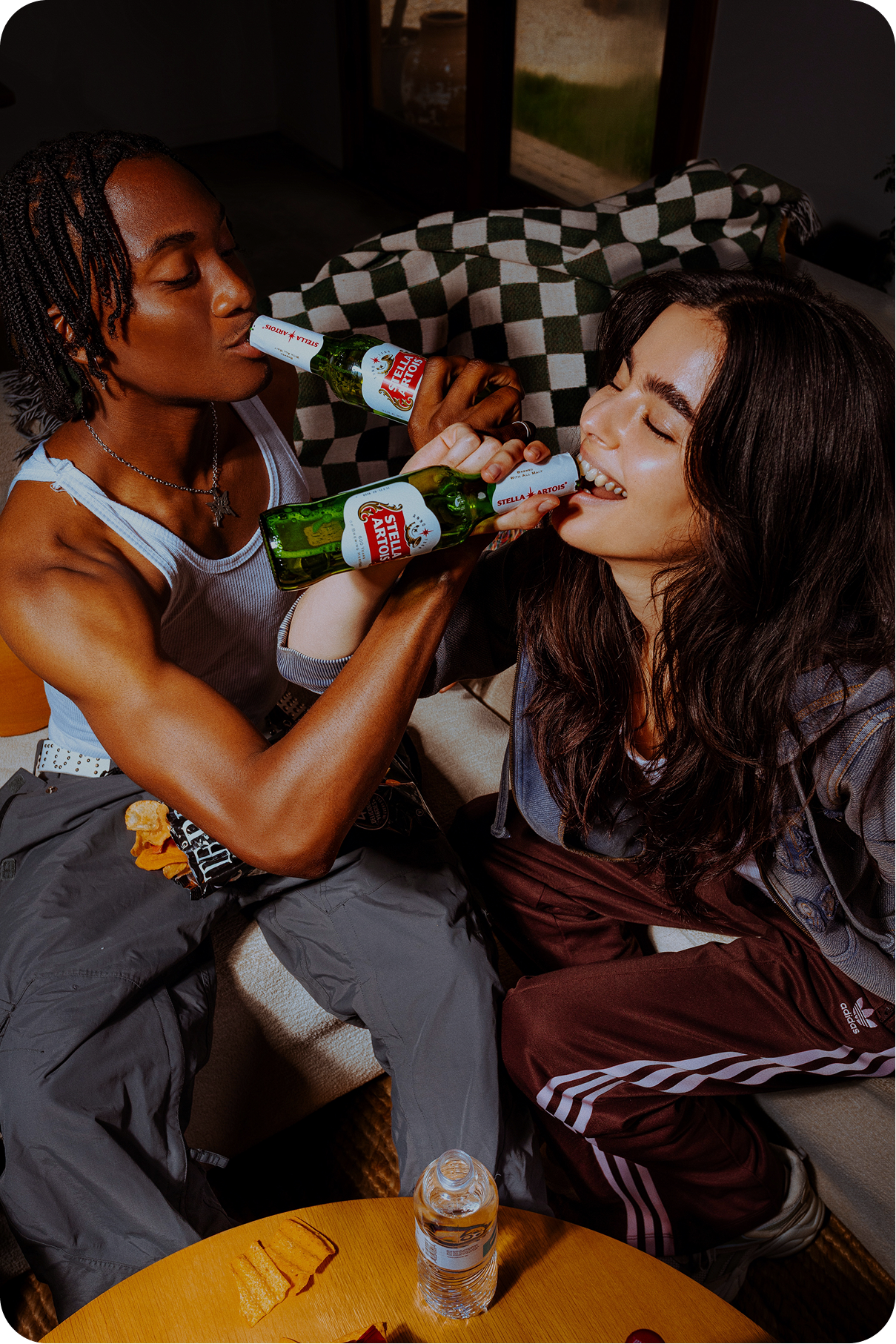 A person with dreadlocks and a white tank top sits on a sofa, drinking from a green bottle of Stella Artois. Another person with long, dark hair sits next to them, also drinking from a Stella Artois bottle. They are both smiling.