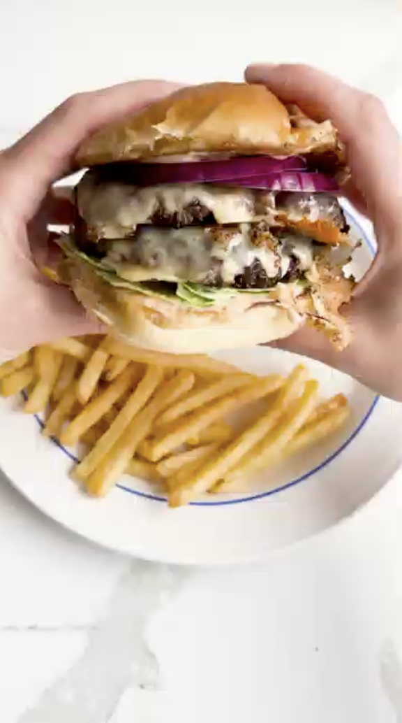 Hands holding a Gopuff double cheeseburger with melted cheese, red onion, and lettuce, served with fries.