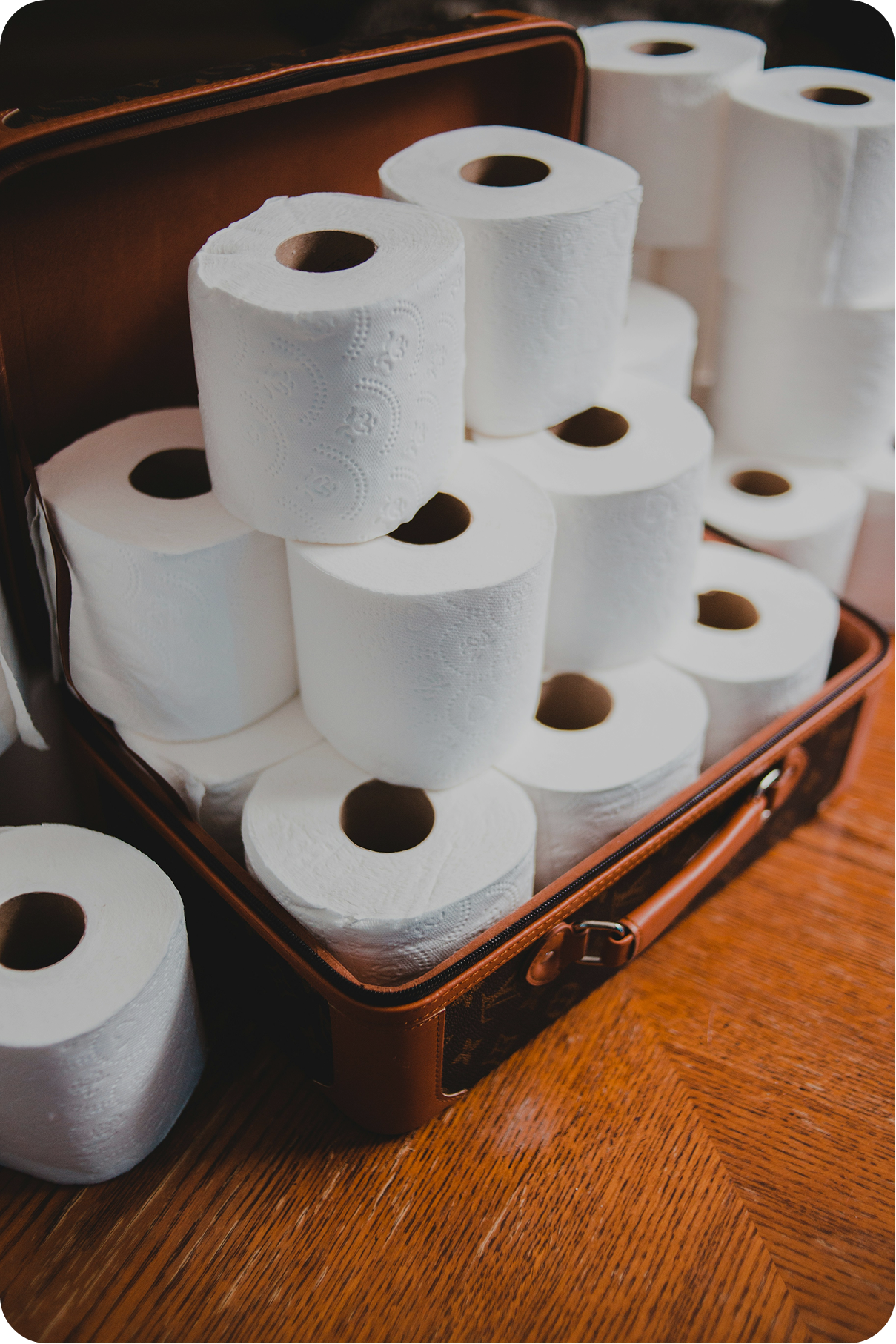 A suitcase filled to the brim with stacks of white toilet paper rolls is open on a wooden floor. More rolls are piled high around the suitcase.